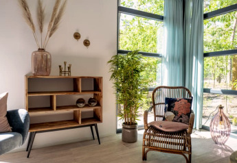 Lodge interieur met rotan stoel, groene planten, houten kast en grote ramen met uitzicht op buiten.
