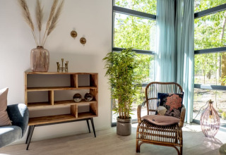 Intérieur de lodge lumineux avec fauteuil en rotin, plantes, étagère en bois et grandes fenêtres vitrées.