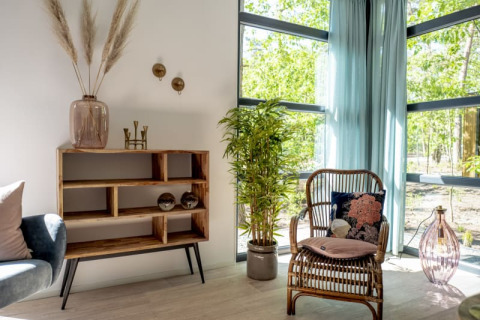 Intérieur de lodge lumineux avec fauteuil en rotin, plantes, étagère en bois et grandes fenêtres vitrées.