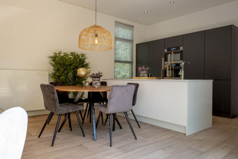 Moderne lodgekøkken med sort skab, hvid ø, spisebord, grå stole, planter og naturlig belysning.