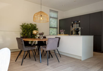 Moderne lodgekøkken med sort skab, hvid ø, spisebord, grå stole, planter og naturlig belysning.