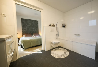 Modern bathroom with wall-mounted toilet, round rug, large bathtub, and doorway leading to bedroom.