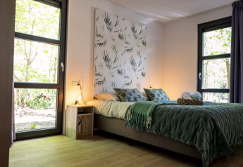 Moderne lodge slaapkamer met grote ramen, groen beddengoed, natuur wanddecoratie en lichte houten vloer.