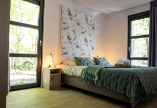 Modern lodge bedroom with large windows, green bed covers, nature wall art, and light wooden flooring.