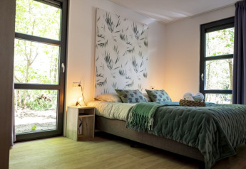 Moderne lodge-soveværelse med store vinduer, grøn sengetæppe og naturmotiv på væggen, lyst trægulv.
