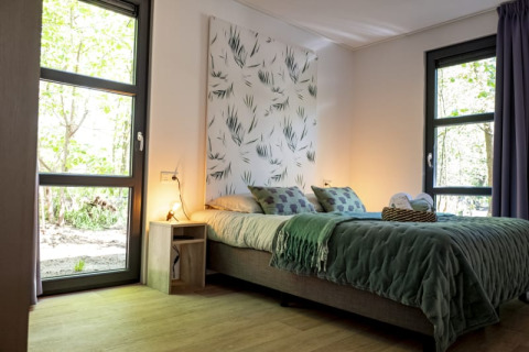 Modern lodge bedroom with large windows, green bed covers, nature wall art, and light wooden flooring.