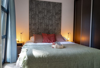 Dormitorio acogedor en una cabaña, con cama grande, manta verde, cojines rosas y luz ambiental suave.