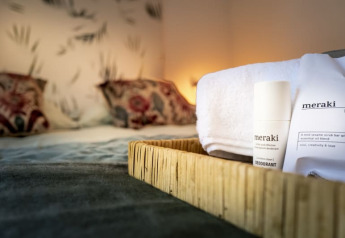 Et nærbillede af en bakke med Meraki toiletartikler og håndklæde på en seng i et hyggeligt lodgeværelse.