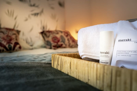 Close-up of a tray with Meraki toiletries and a towel on a bed in a cozy lodge room with soft lighting.