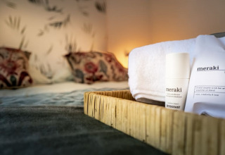 Close-up of a tray with Meraki toiletries and a towel on a bed in a cozy lodge room with soft lighting.