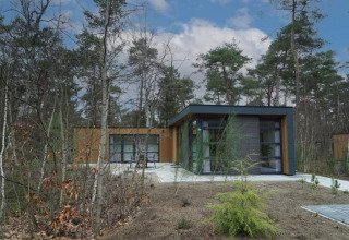 Lodge moderne avec de grandes fenêtres, entouré de forêt et de nature, vu de l’extérieur par temps ensoleillé.