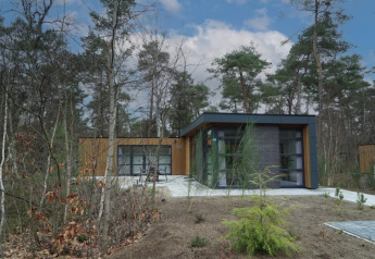 Moderne lodge med store vinduer omgivet af skov og natur, set udefra på en solrig dag med blå himmel.