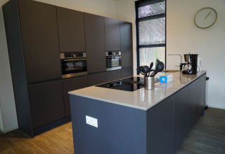 Cocina moderna en una cabaña, con muebles oscuros, isla central, cafetera y ventanas grandes.