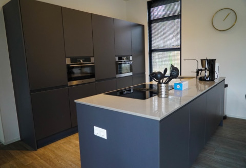 Cocina moderna en una cabaña, con muebles oscuros, isla central, cafetera y ventanas grandes.