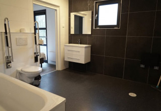 Baño accesible en una cabaña con sanitarios modernos, paredes de azulejo negro y ventana grande.