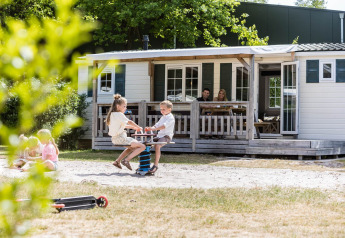 Bambini giocano fuori da uno chalet familiare mentre gli adulti si rilassano in veranda a Kampeerdorp de Zandstuve.