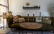 Hyggelig lodge-stue med grøn sofa, mønstrede puder, to tændte stearinlys og planter i fletkurve.