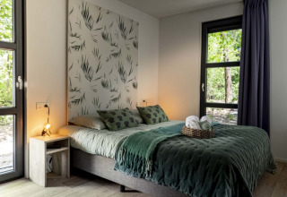 Dormitorio moderno en una cabaña con grandes ventanas, ropa de cama verde y vista a la naturaleza.