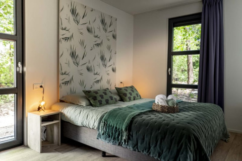 Dormitorio moderno en una cabaña con grandes ventanas, ropa de cama verde y vista a la naturaleza.