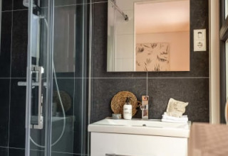Baño moderno en lodge con ducha de vidrio, baldosas oscuras, mueble blanco y espejo sobre el lavabo.