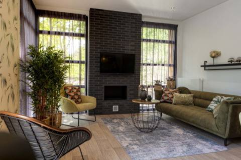 Sala moderna de lodge con sofá verde, sillón, chimenea, grandes ventanales y plantas decorativas.