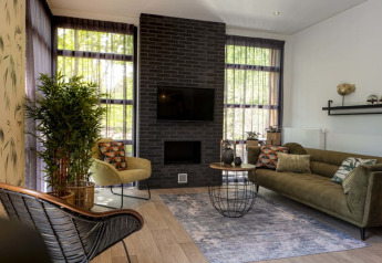 Salon moderne de lodge avec canapé vert, fauteuil, cheminée, grandes fenêtres et plantes vertes décoratives.