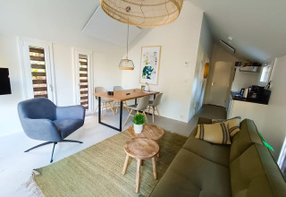 Modernes Lodge-Wohnzimmer mit grünem Sofa, grauem Sessel, Holztischen, Esstisch und viel Tageslicht.