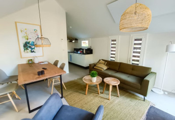Intérieur de lodge moderne avec salon ouvert, canapé vert, coin repas, tables en bois et lampes en osier.