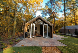 Photo d’un lodge moderne en pleine forêt avec portes doubles ouvertes et feuilles d’automne au sol.