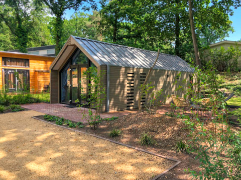 Un lodge moderne au toit métallique, entouré d’arbres et de chemins de jardin, avec des sièges en plein air.