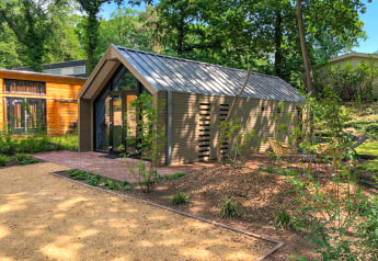 Un lodge moderno con tetto in metallo, immerso nel verde, circondato da sentieri e sedute da giardino.