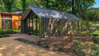 Un lodge moderne au toit métallique, entouré d’arbres et de chemins de jardin, avec des sièges en plein air.