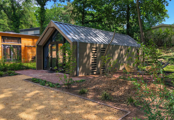 Un albergue moderno con techo de metal, rodeado de árboles y senderos de jardín, con asientos exteriores.