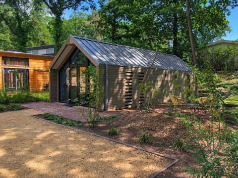Un albergue moderno con techo de metal, rodeado de árboles y senderos de jardín, con asientos exteriores.