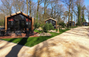 Photo of modern wooden lodges along a sandy path, surrounded by trees and greenery in a forest setting.