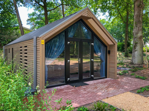Moderne houten lodge met grote glazen deuren en blauwe gordijnen, omgeven door groen en bomen buiten.