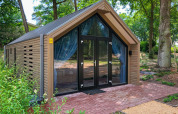 Lodge moderne en bois avec grandes portes vitrées et rideaux bleus, entouré de verdure et d’arbres en extérieur.