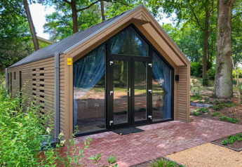 Modern wooden lodge house with large glass doors and blue curtains, surrounded by greenery and trees outdoors.