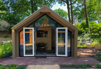 Et billede af en hyggelig lodge med store åbne døre, omgivet af natur og grønne træer i baggrunden.