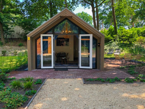 Modern lodge with glass doors, brick patio, and outdoor seating, surrounded by lush green forest.