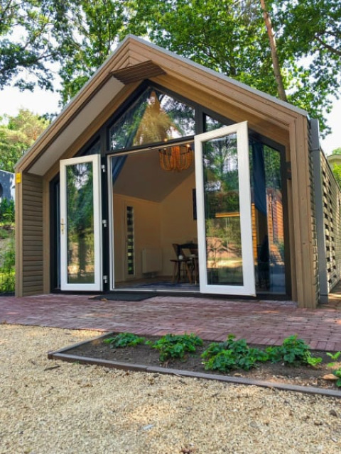 Lodge moderne avec portes vitrées ouvertes, bois et petit jardin, entouré de grands arbres verdoyants.