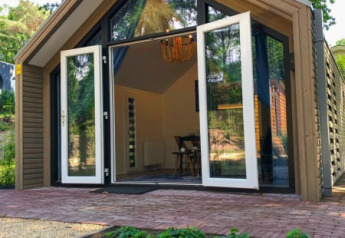 Moderne lodge med åbne glasdøre og træbeklædning, omgivet af grønne træer og en lille have foran.