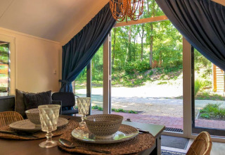 Comedor en una cabaña con vajilla elegante, puertas de cristal abiertas y vistas a un frondoso bosque verde afuera.