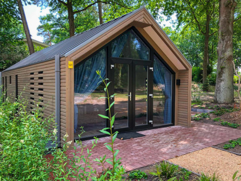 Moderne houten lodge met grote glazen deuren, omgeven door planten en bomen in een bosrijke omgeving.