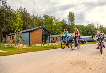 Drie mensen fietsen langs moderne lodges die te midden van groen en onder een bewolkte hemel liggen.