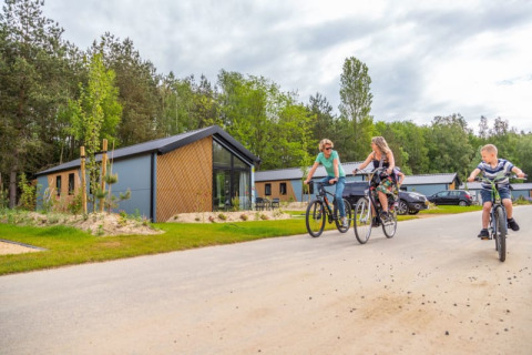 Drie mensen fietsen langs moderne lodges die te midden van groen en onder een bewolkte hemel liggen.