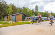 Drei Menschen fahren Fahrrad vor modernen Lodges, umgeben von Bäumen und parkenden Autos unter Wolken.