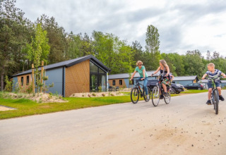 Drie mensen fietsen langs moderne lodges die te midden van groen en onder een bewolkte hemel liggen.