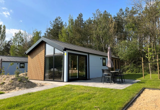 Moderne Lodge mit großen Fenstern und Terrasse im Grünen, umgeben von Bäumen und gepflegtem Rasen.