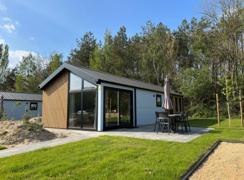 Moderne lodge met grote ramen, terras met tuinset en parasol, omringd door gras en bomen in het groen.